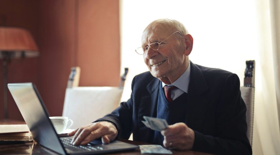 An elderly man is sitting at a table with his right hand using a laptop and his left hand holding cash. He has a smile on his face and his reading glasses on.